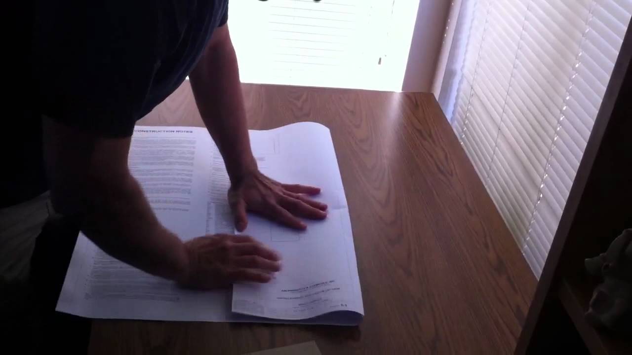 A mans hands folding up blueprints on top of a desk near some windows.