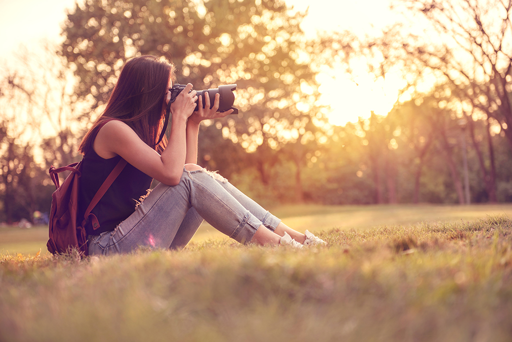 A woman in profile seated in grass late in the day, holding a camera up to her face.