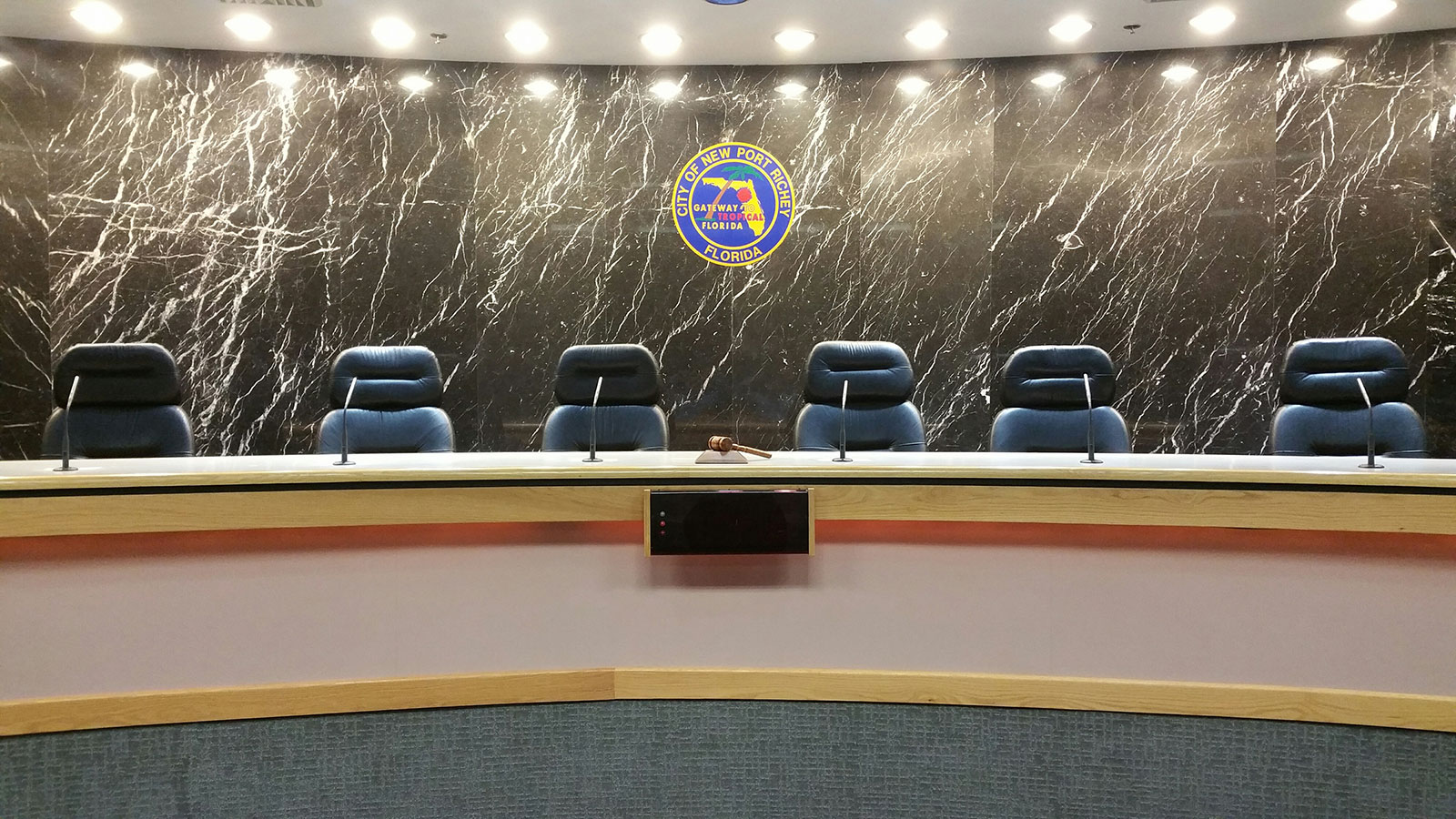 A dais with microphones mounted in it, chairs are set up behind it. The City of New Port Richey seal is visible behind them