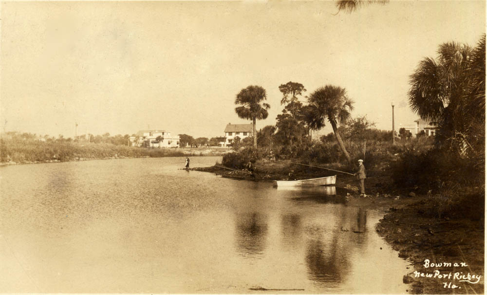 Sepia toned riverbank with man fishing next to a small boat. Houses visible in background.