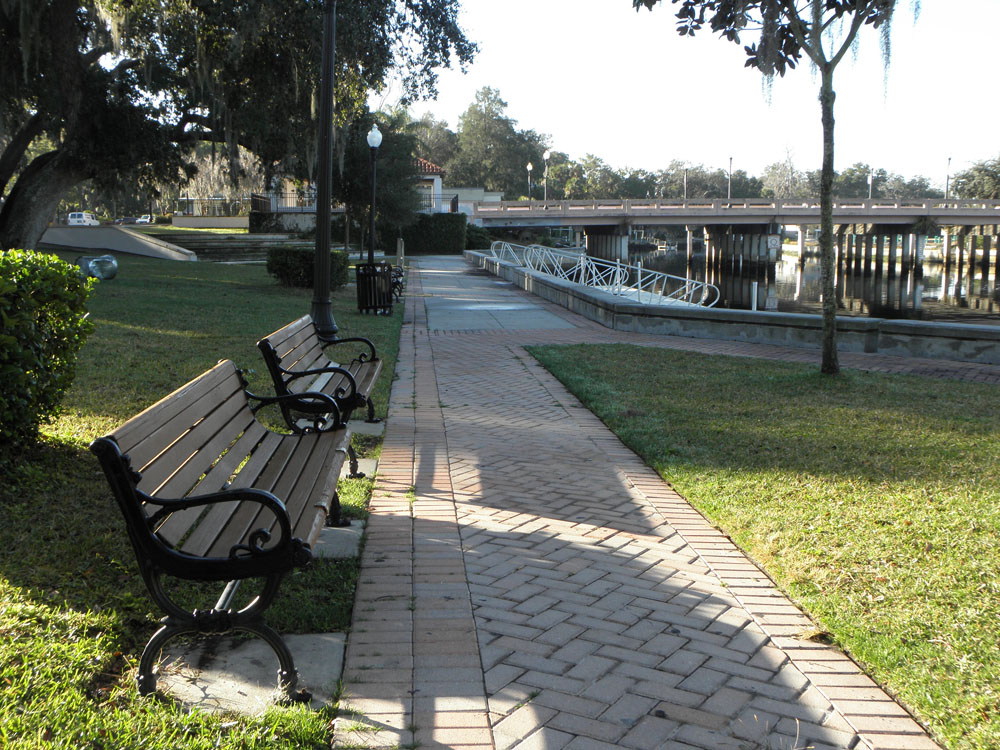 A brick sidewalk in a park with benches along it. A bridge over a river is visible in the background