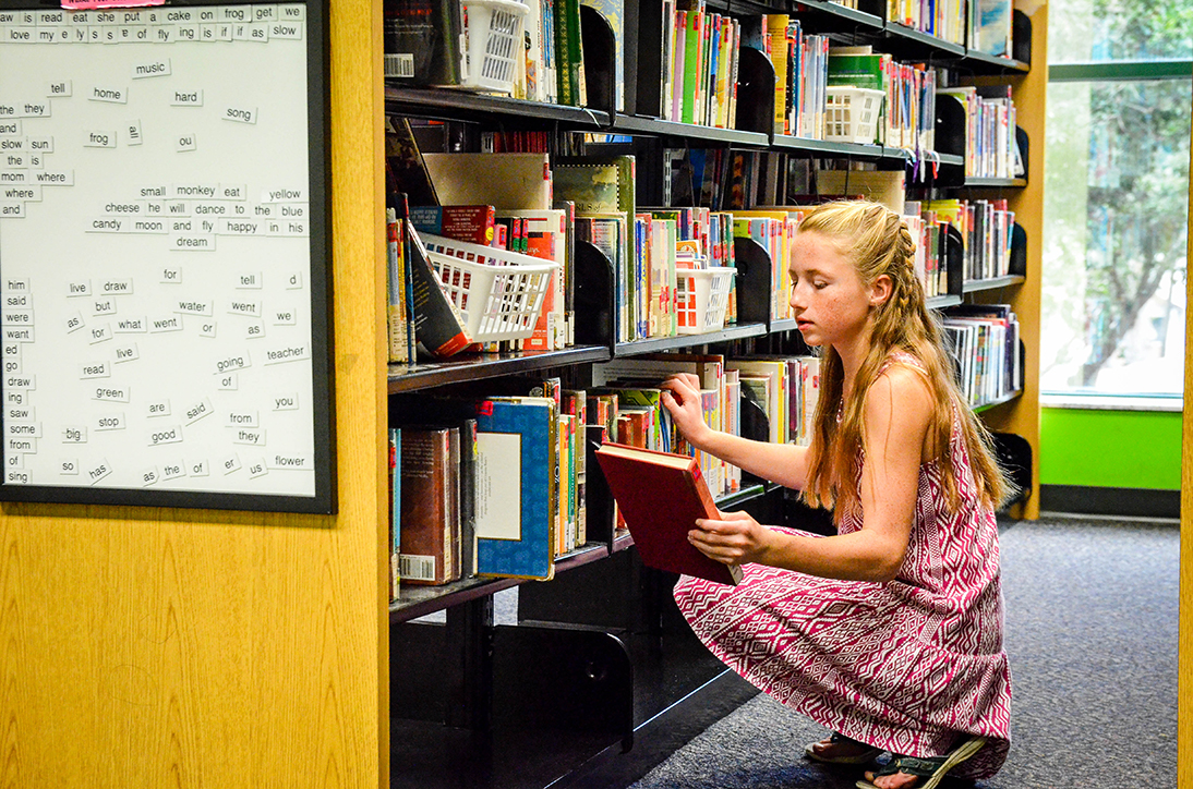 A young girl with light colored hair squatting in front of a bookshelf at a library. She is holding a red book.