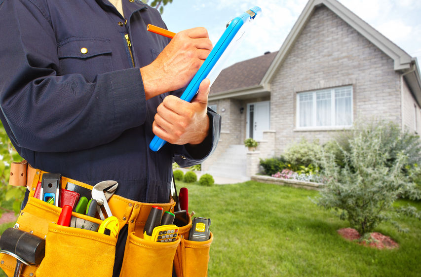 A man writing in a blue clip board wearing a yellow utility belt full of tools standing outside of a house