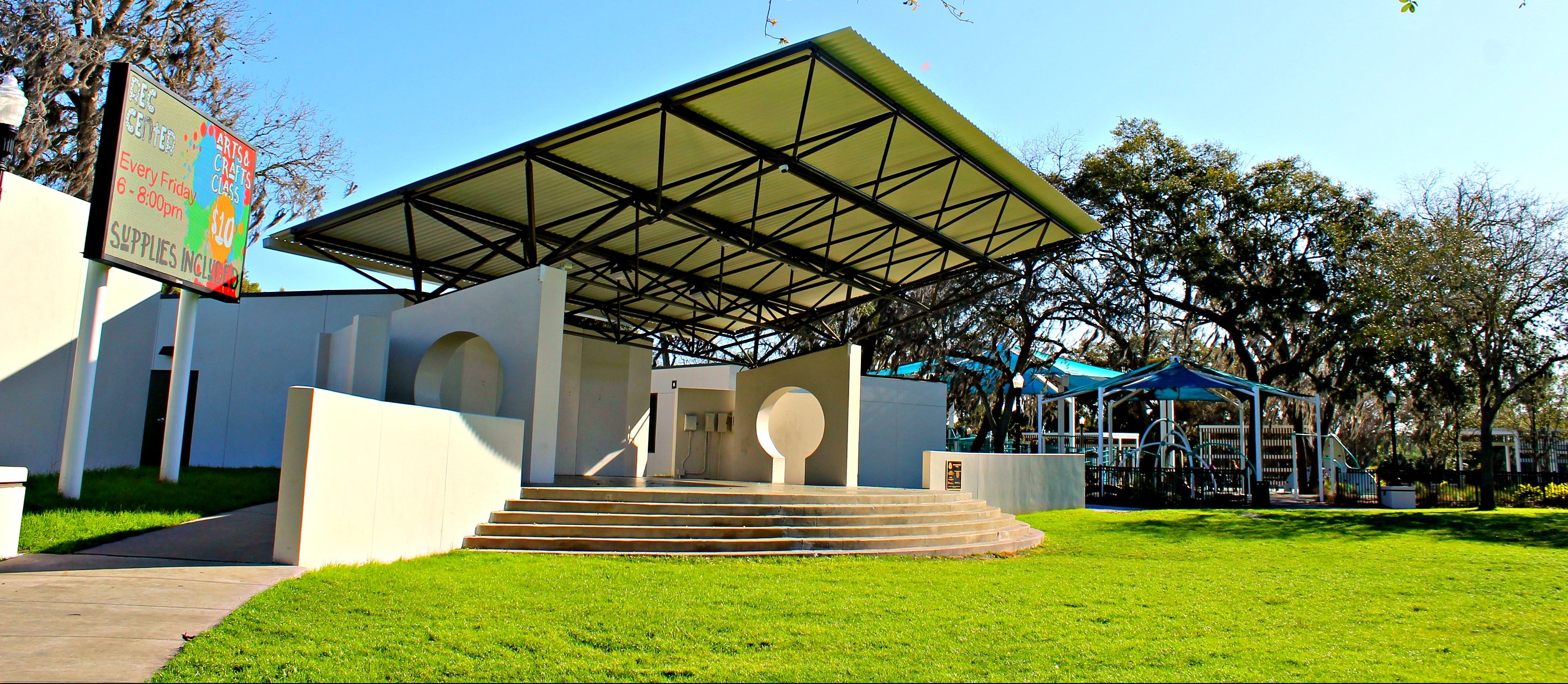 An outdoor curved concrete thrust stage with an awning and wraparound stairs in a sunny field. A playground is visible in the background.