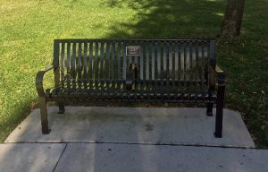 An iron bench with a dedication plaque and an arm rest in the middle.