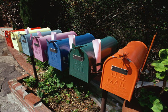 A row of identically shaped mailboxes of varying colors, planted in a raised brick flower bed.