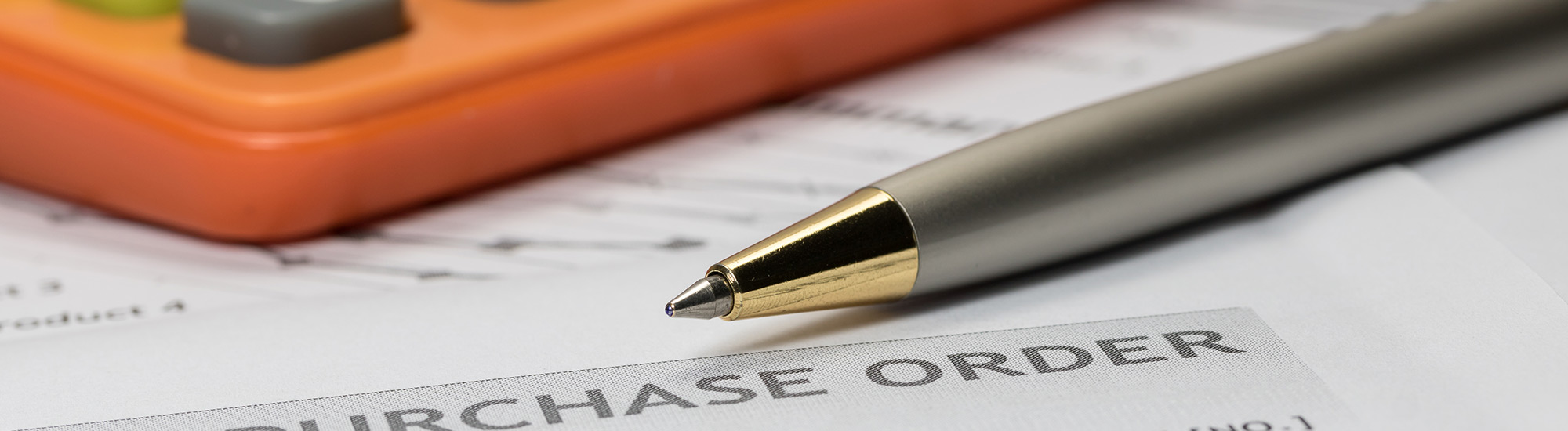 Closeup of the tip of a pen laying on some papers. the letters "U R C H A S E O R D E R" are visible on the paper. The edge of an orange calculator is visible in the background.