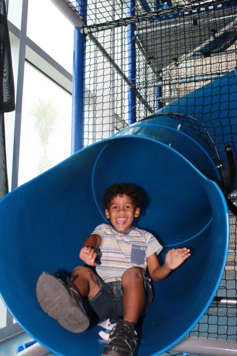 a young boy wearing a multi colored stripped shirt, smiling while reaching the end of a large blue slide.